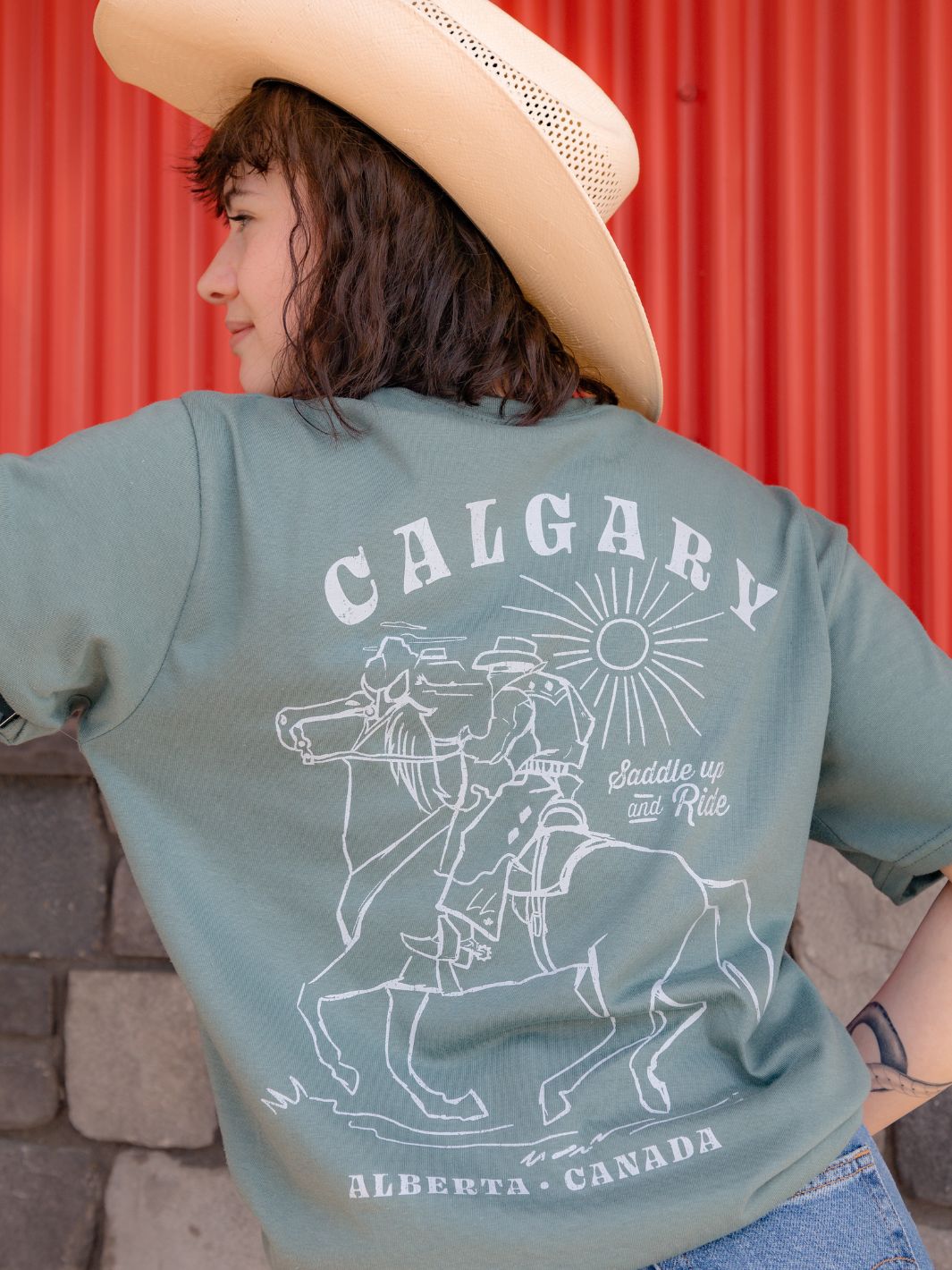 Individual showcasing the Local Laundry Saddle Up and Ride shirt in Sage green featuring the back graphic of a cowboy riding a horse with 