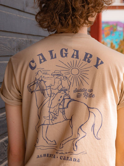 Individual wearing Saddle Up and Ride shirt showcasing a back graphic of a cowboy riding a horse into the sunset with Calgary spelled out across the top and Alberta, Canada across the bottom of the graphic. Made in Canada - Local Laundry