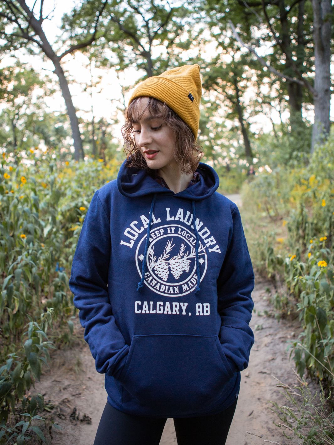 Individual hiking in the woods donning their Local Laundry Varsity hoodie in Navy blue, featuring a white graphic on the front torso of the hoodie. Individual is pairing this hoodie with the Local Laundry Giving Toque in Mustard.