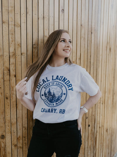 Happy individual wearing the Local Laundry Varsity t-shirt. This shirt features a contrasting blue crest graphic featuring a forest scape with the words "Keep it Local" on the top, and "Canadian Made" across the bottom, with "Local Laundry" overarching atop of the crest.