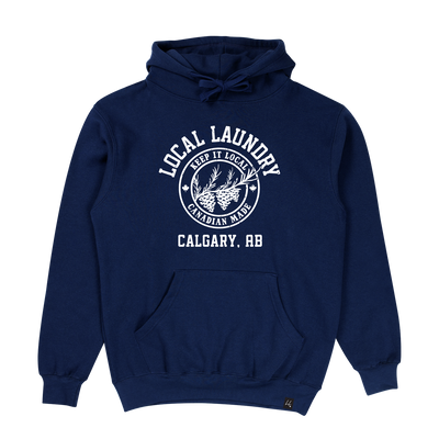 The Local Laundry Varsity hoodie in navy blue featuring a white graphic on the front torso. The graphic features a crest with three pine cones and "Keep it Local" on the top and "Canadian Made" on the bottom. This garment is sustainably made in Canada, utilizing a premium blend of 50/50 cotton and polyester.