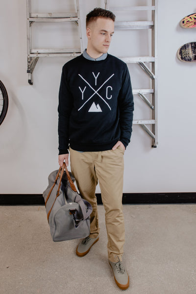 Individual showcasing their YYC Crewneck sweater with tan trousers and a grey and brown duffle bag. Made in Canada, ethically and sustainably.
